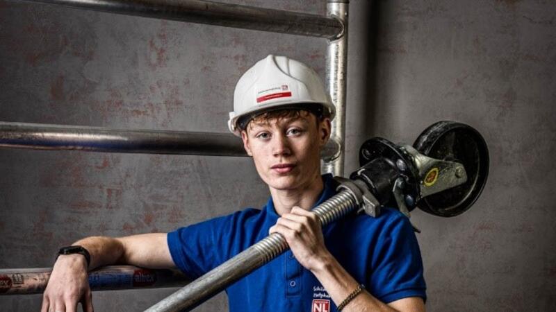 Een jongeman in werkkleding staat stoer bij een steiger en heeft een steigeronderdeel in zijn hand