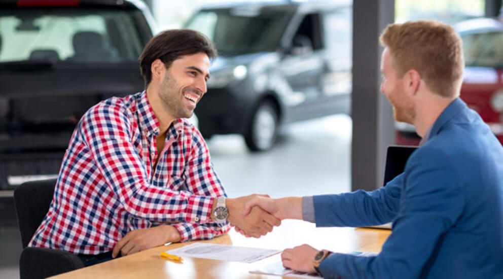 Twee mannen zitten aan  een tafel bij een autodealer en ze schudden elkaar lachend de hand